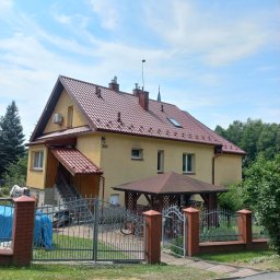 LUXPURE - Dwupiętrowy dom z nowym, błyszczącym, czerwonym dachem z dachówki ceramicznej, widoczny również fragment rynny, żółta elewacja z numerem 303, metalowe ogrodzenie i altana w ogrodzie.