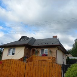 LUXPURE - Dwupiętrowy dom z ciemnoszarym dachem z blachy trapezowej, widok od strony drewnianego ogrodzenia i zieleni ogrodu, pochmurne niebo w tle.