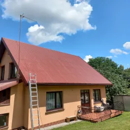 Dom jednorodzinny z nowym, czerwonym dachem z blachy trapezowej, drabina oparta o ścianę, antena telewizyjna na dachu, balkon z kutą balustradą, błękitne niebo z białymi chmurami.