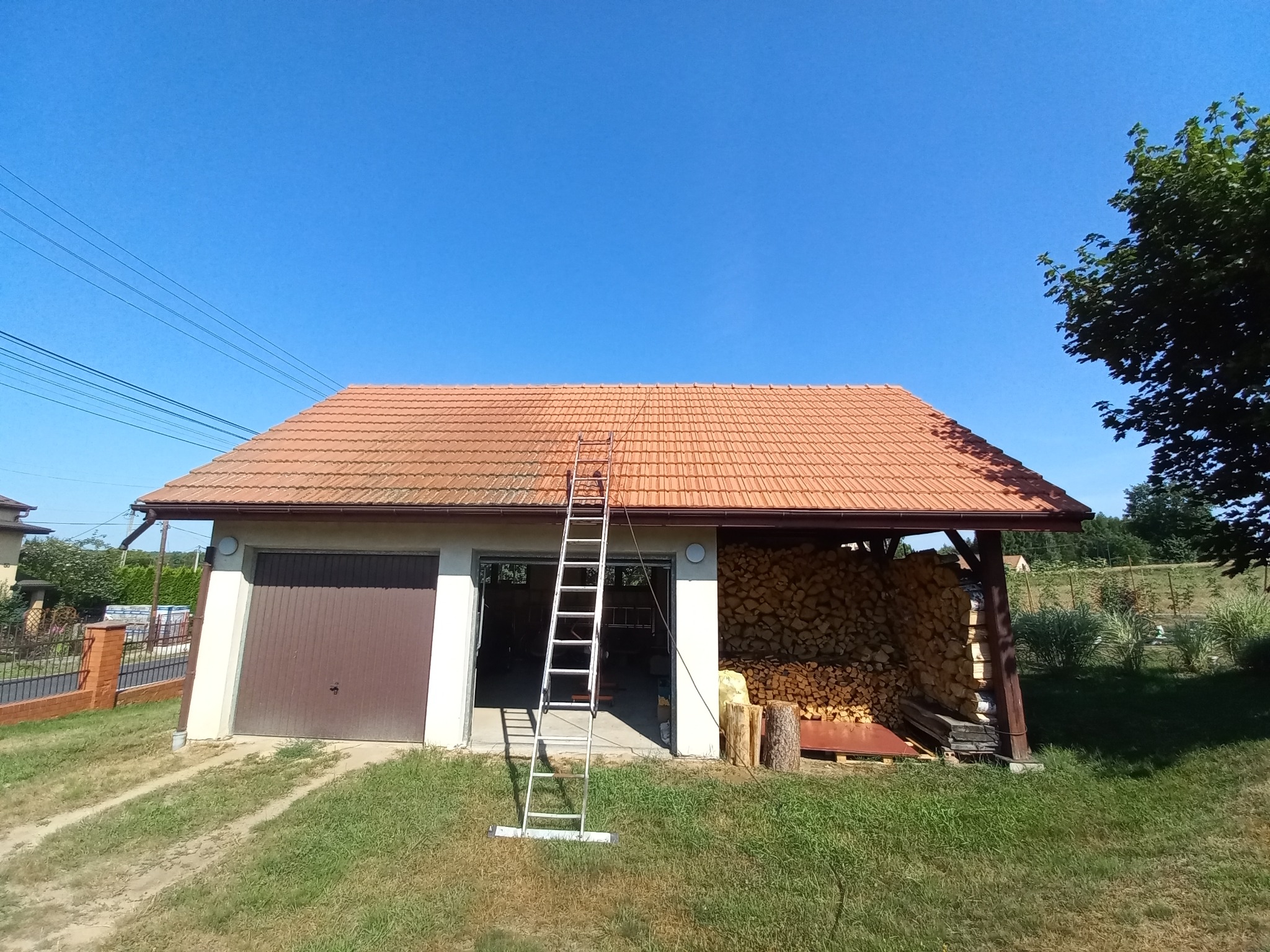 Częściowo wyczyszczony, pomarańczowy dach z dachówki ceramicznej, drabina oparta o budynek gospodarczy z otwartym garażem i składowanym drewnem, widoczne ślady czyszczenia.