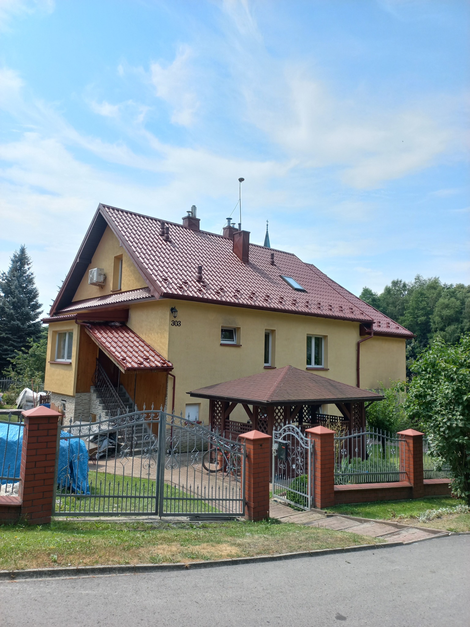 Dwupiętrowy dom z nowym, błyszczącym, czerwonym dachem z dachówki ceramicznej, widoczny również fragment rynny, żółta elewacja z numerem 303, metalowe ogrodzenie i altana w ogrodzie.