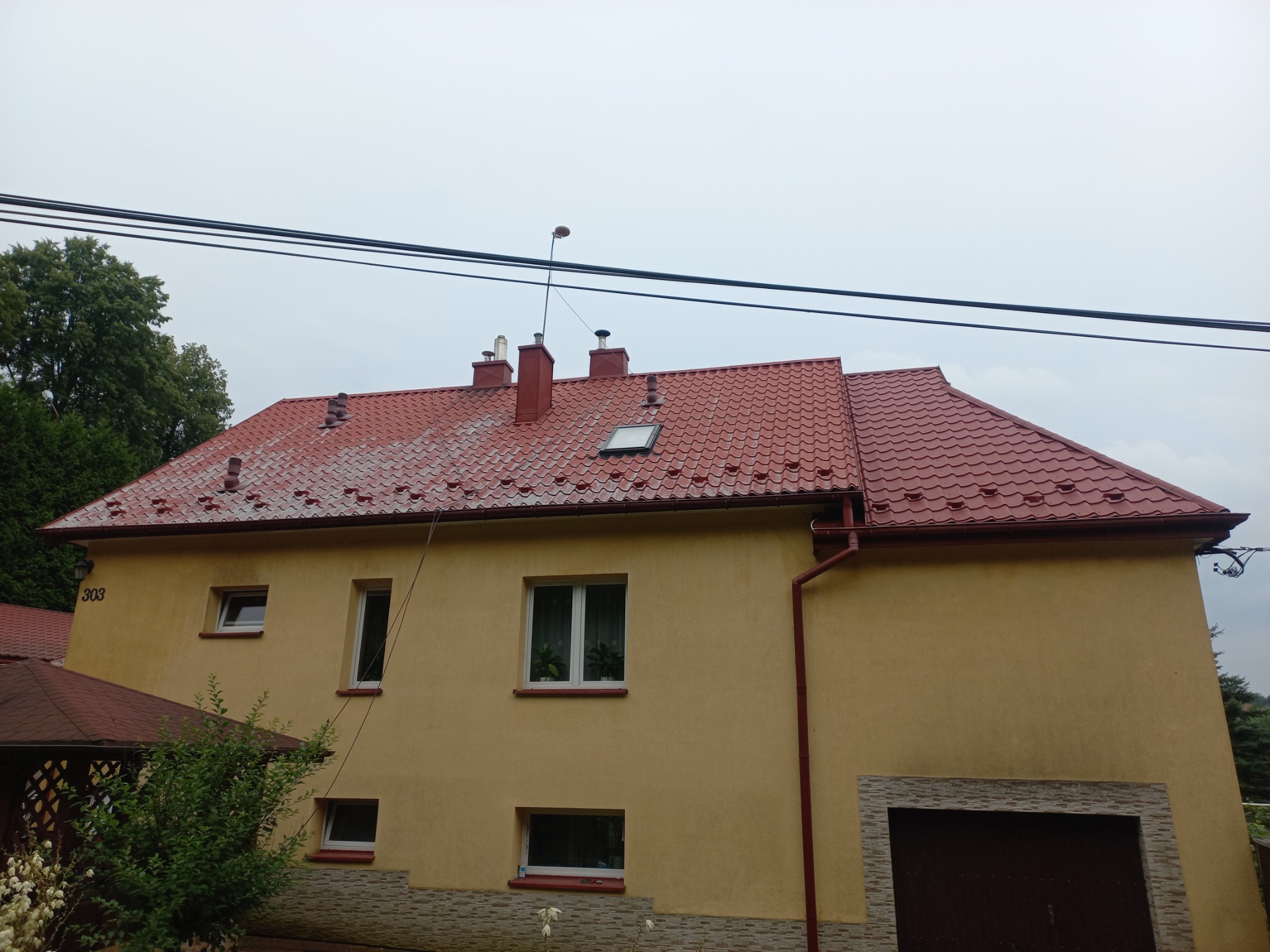 Dwupiętrowy dom z nowym, czerwonym dachem z dachówki ceramicznej, widoczne trzy kominy, okno dachowe oraz rynny w kolorze dachu. Część dachu wykazuje ślady zabrudzeń.