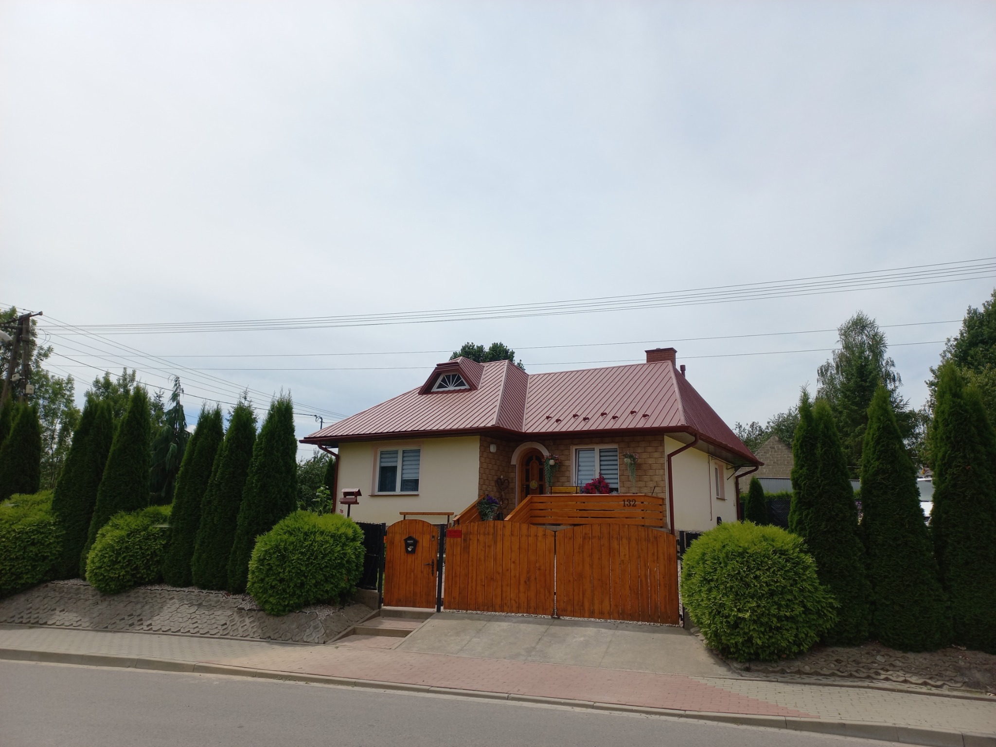 Dom jednorodzinny z czerwonym dachem dwuspadowym, drewnianym ogrodzeniem i zadbanym ogrodem, widok z ulicy w pochmurny dzień.