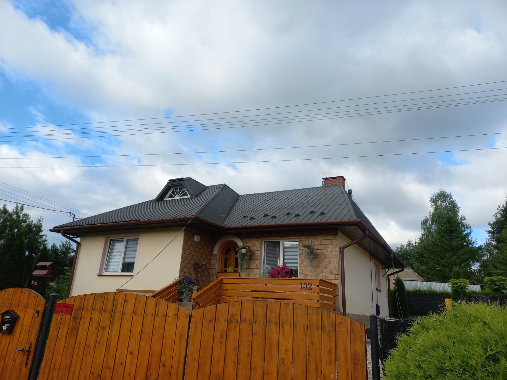 Dwupiętrowy dom z ciemnoszarym dachem z blachy trapezowej, widok od strony drewnianego ogrodzenia i zieleni ogrodu, pochmurne niebo w tle.