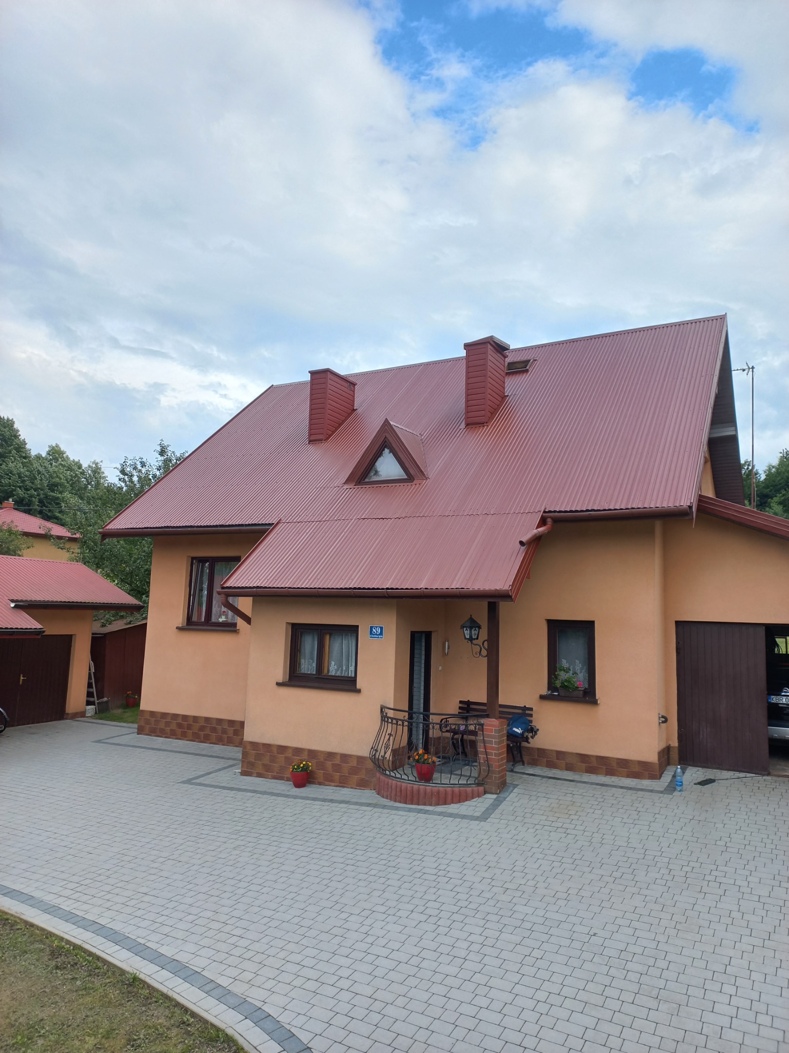 Dom jednorodzinny z nowym, brązowo-czerwonym dachem z blachy trapezowej, widoczne kominy, rynny i okno dachowe w kształcie trójkąta. Na elewacji numer domu '89'.