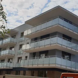 Nowoczesny budynek wielorodzinny z balkonami zabezpieczonymi szklanymi balustradami, widok z dołu na tle błękitnego nieba z chmurami, z widocznym fragmentem drzewa w lewym górnym rogu i fragmentem...