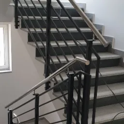 Wewnętrzne schody z szarymi, granitowymi stopniami i czarną, metalową balustradą z poziomymi prętami oraz srebrnymi poręczami, widok z dołu.