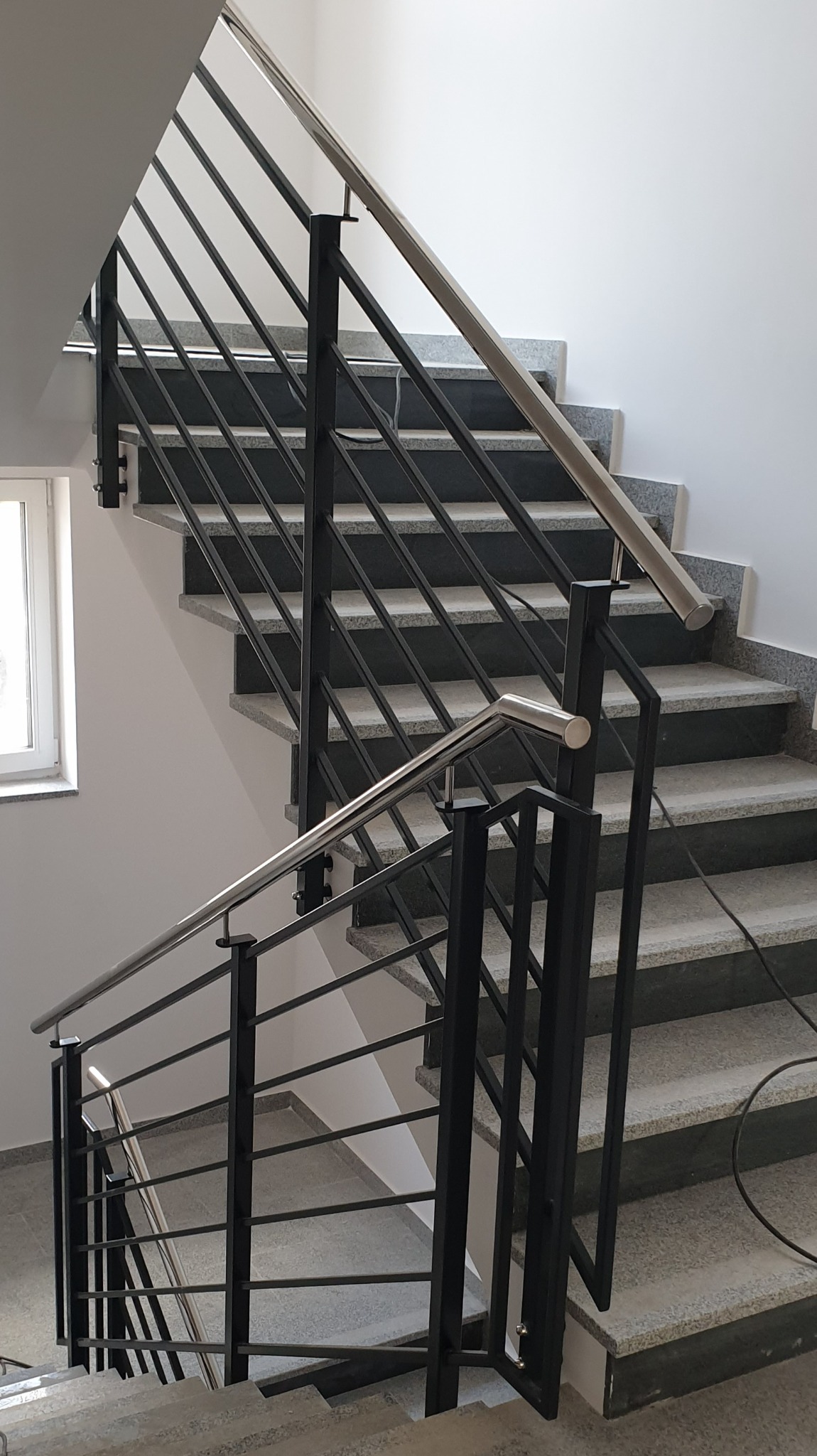 Wewnętrzne schody z szarymi, granitowymi stopniami i czarną, metalową balustradą z poziomymi prętami oraz srebrnymi poręczami, widok z dołu.