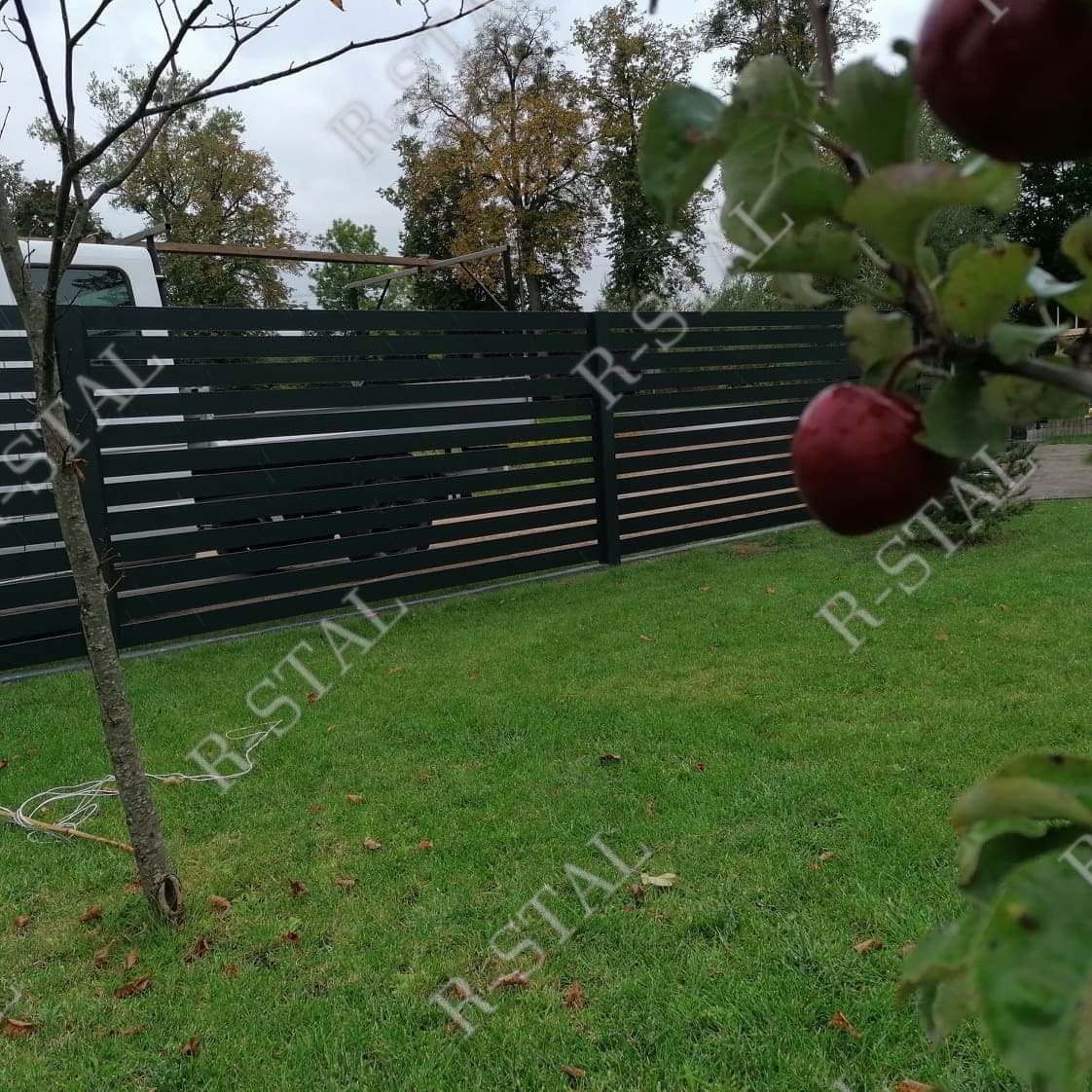 Nowoczesne ogrodzenie panelowe z poziomych listew w kolorze antracytowym, z widocznym fragmentem białego busa w tle i jabłkami na drzewie w pierwszym planie.