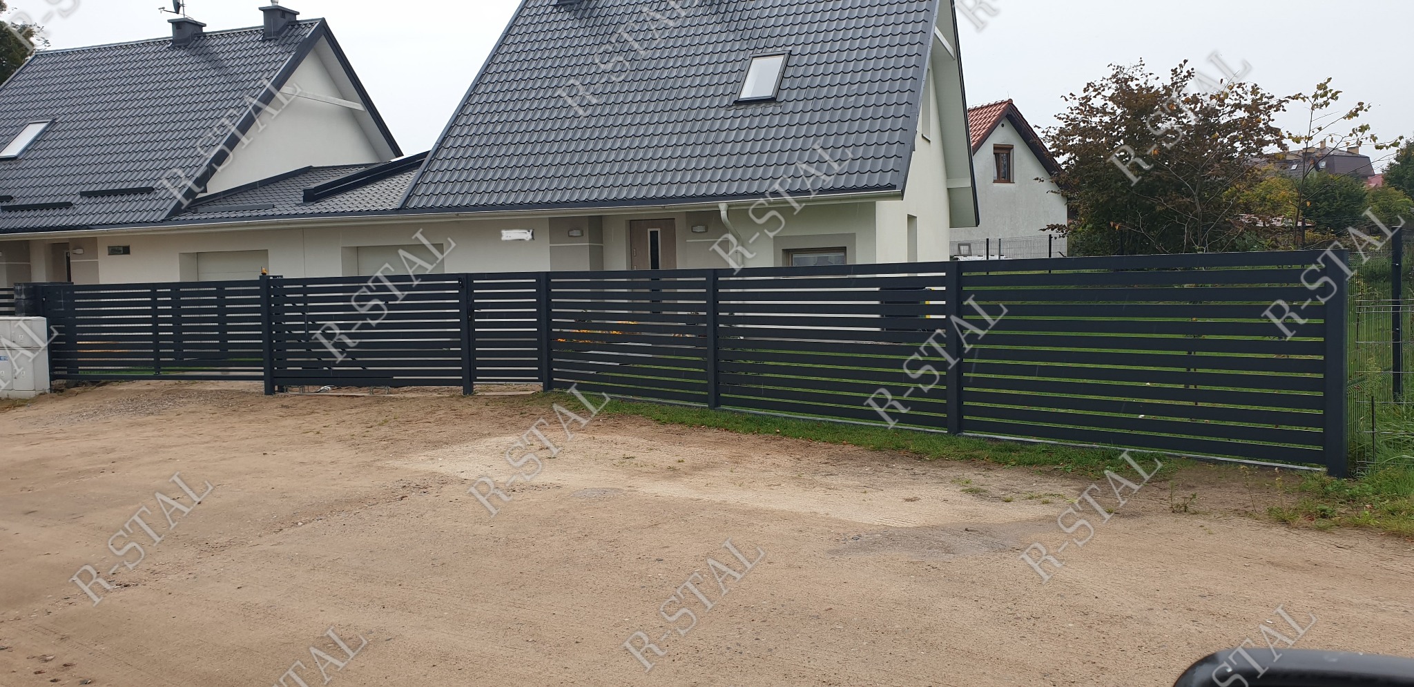 Nowoczesne ogrodzenie panelowe z poziomych profili w kolorze antracytowym, zamontowane przed domem jednorodzinnym z szarą dachówką, widoczne z poziomu gruntu.