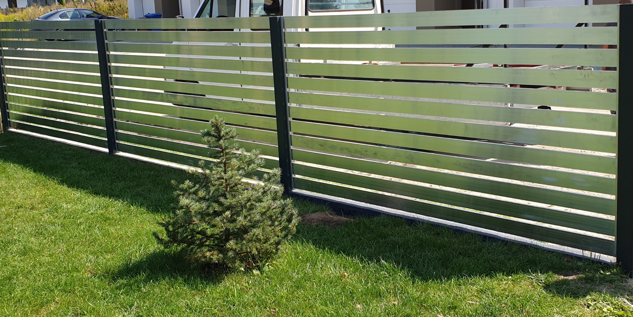 Nowoczesne ogrodzenie z poziomych, metalowych paneli w kolorze jasnego aluminium, z ciemnymi słupkami, na tle zielonego trawnika z małą choinką.