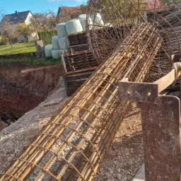 Szkielet zbrojeniowy w kształcie walca i kuli, leżący na żwirowym podłożu obok wykopu, z widocznymi elementami konstrukcyjnymi i stosem materiałów budowlanych w tle.