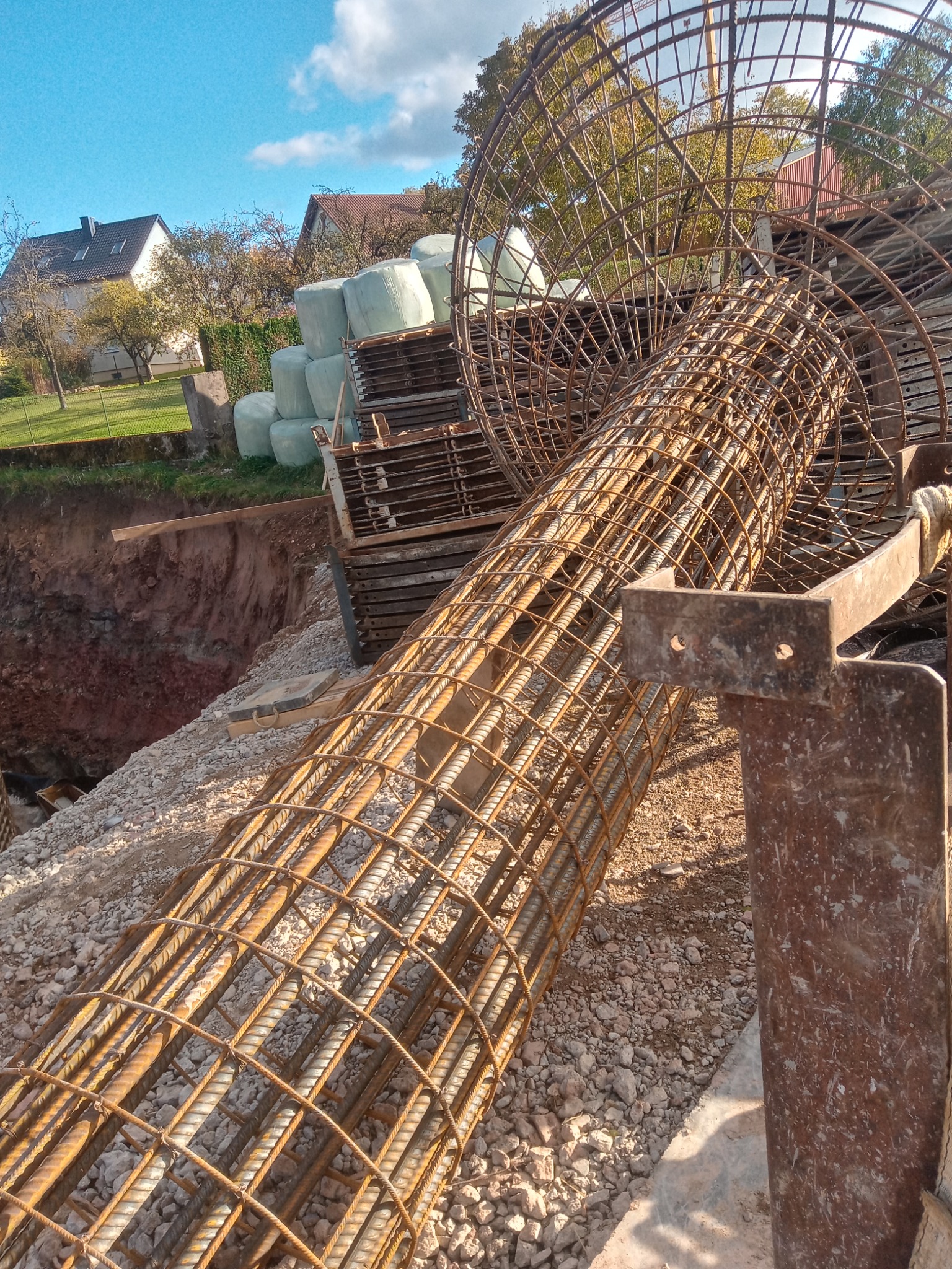 Szkielet zbrojeniowy w kształcie walca i kuli, leżący na żwirowym podłożu obok wykopu, z widocznymi elementami konstrukcyjnymi i stosem materiałów budowlanych w tle.