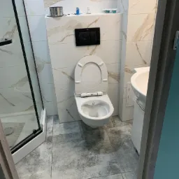 Nowoczesna łazienka z kabiną prysznicową, podwieszaną toaletą z czarnym panelem sterującym i umywalką na szafce, wykończona płytkami imitującymi marmur z brązowymi żyłami i szarymi płytkami...