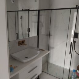 Bodzio. Remonty - Nowoczesna łazienka z kabiną prysznicową typu walk-in z czarnymi ramami, białą umywalką nablatową na szafce z szufladami i złotą armaturą pod lustrem.
