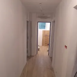 Korytarz w trakcie malowania na biało, z widoczną instalacją elektryczną na suficie i fragmentem wykończonej łazienki w tle.