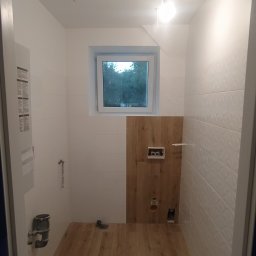 Bodzio. Remonty - Wnętrze łazienki w trakcie remontu: białe płytki na ścianach, drewnopodobne panele na podłodze i częściowo na ścianie, widoczne instalacje wodno-kanalizacyjne, okno z widokiem na zieleń.