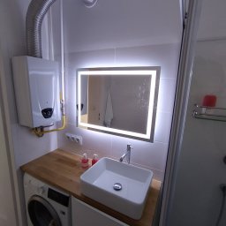 Bodzio. Remonty - Nowoczesna łazienka z podświetlanym lustrem, białą umywalką nablatową, drewnianym blatem nad pralką i gazowym podgrzewaczem wody.