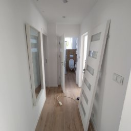 Bodzio. Remonty - Korytarz w Kielcach po remoncie, z drewnianą podłogą, białymi ścianami i nowymi drzwiami z przeszkleniami, widok na odnowioną łazienkę.