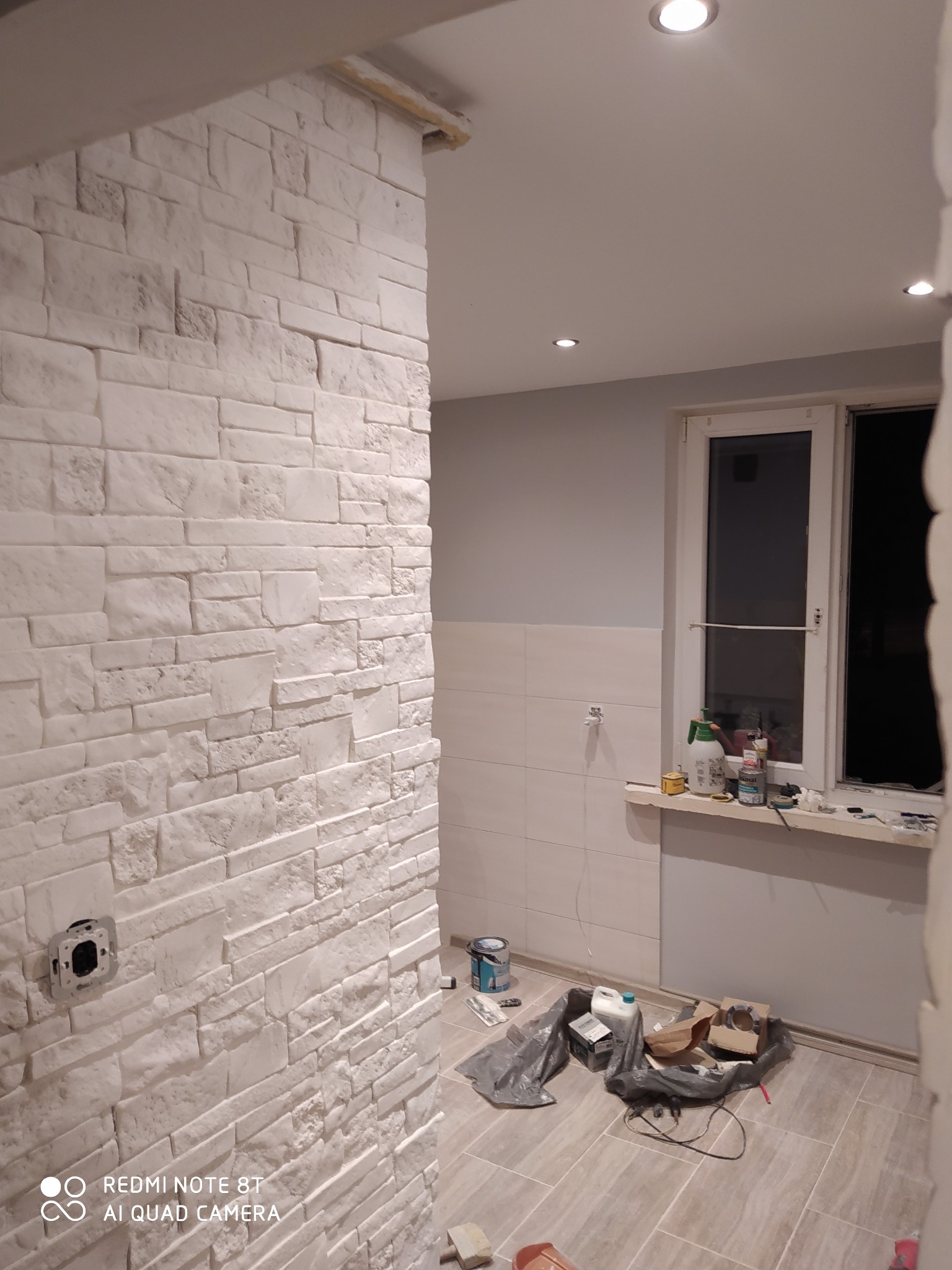 Wykończona ściana z białej cegły dekoracyjnej w trakcie remontu łazienki, widoczne narzędzia i materiały budowlane na podłodze z płytek, gniazdko elektryczne bez osprzętu.