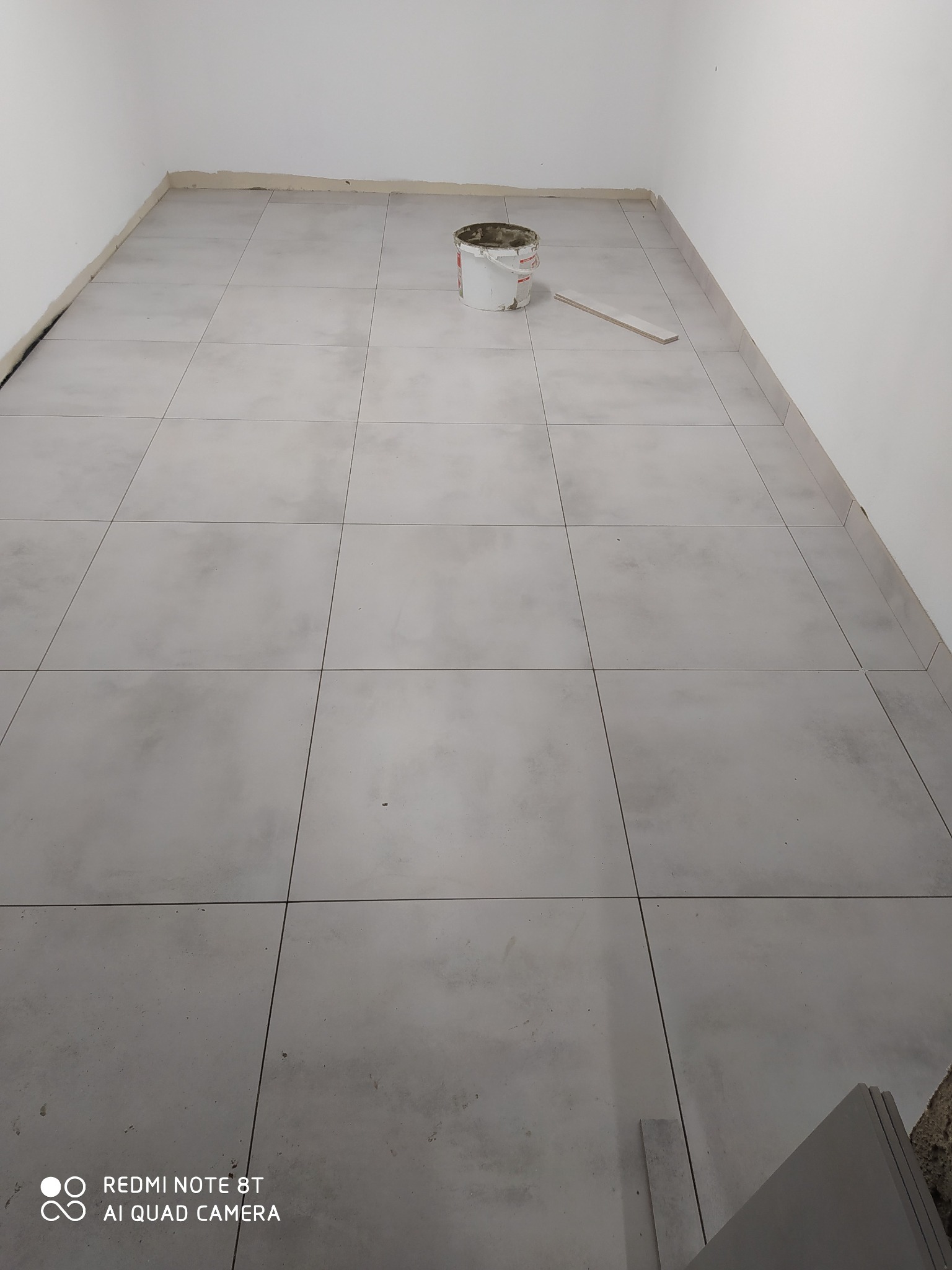 Szare, matowe płytki podłogowe w trakcie układania, widoczny biały kubeł z zaprawą i fragment listwy przypodłogowej.