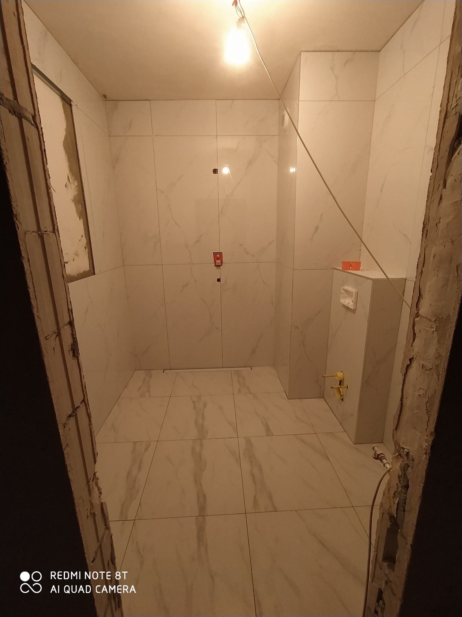 Wnętrze łazienki w trakcie remontu, widoczne białe płytki imitujące marmur na ścianach i podłodze, częściowo wykończone obramowanie drzwi, zamontowana podtynkowa spłuczka WC z wystającymi rurami.