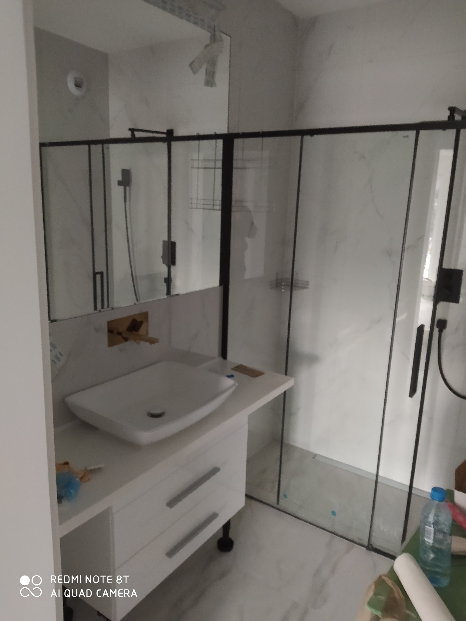 Nowoczesna łazienka z kabiną prysznicową typu walk-in z czarnymi ramami, białą umywalką nablatową na szafce z szufladami i złotą armaturą pod lustrem.