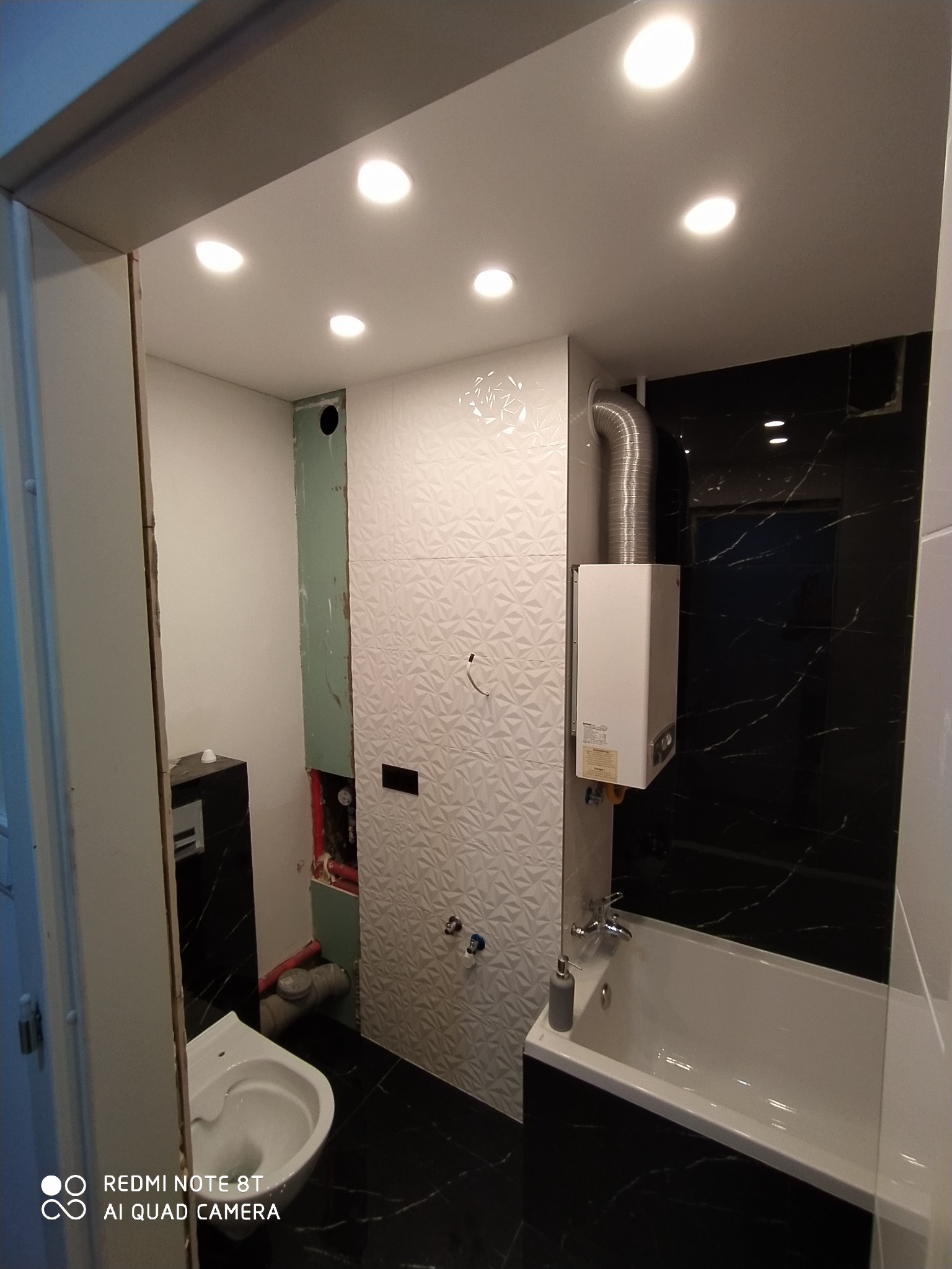 Łazienka w trakcie remontu: widoczne płytki strukturalne na ścianie, czarne płytki imitujące marmur na podłodze i ścianie prysznicowej, częściowo zabudowany stelaż podtynkowy WC, bojler z rurą...