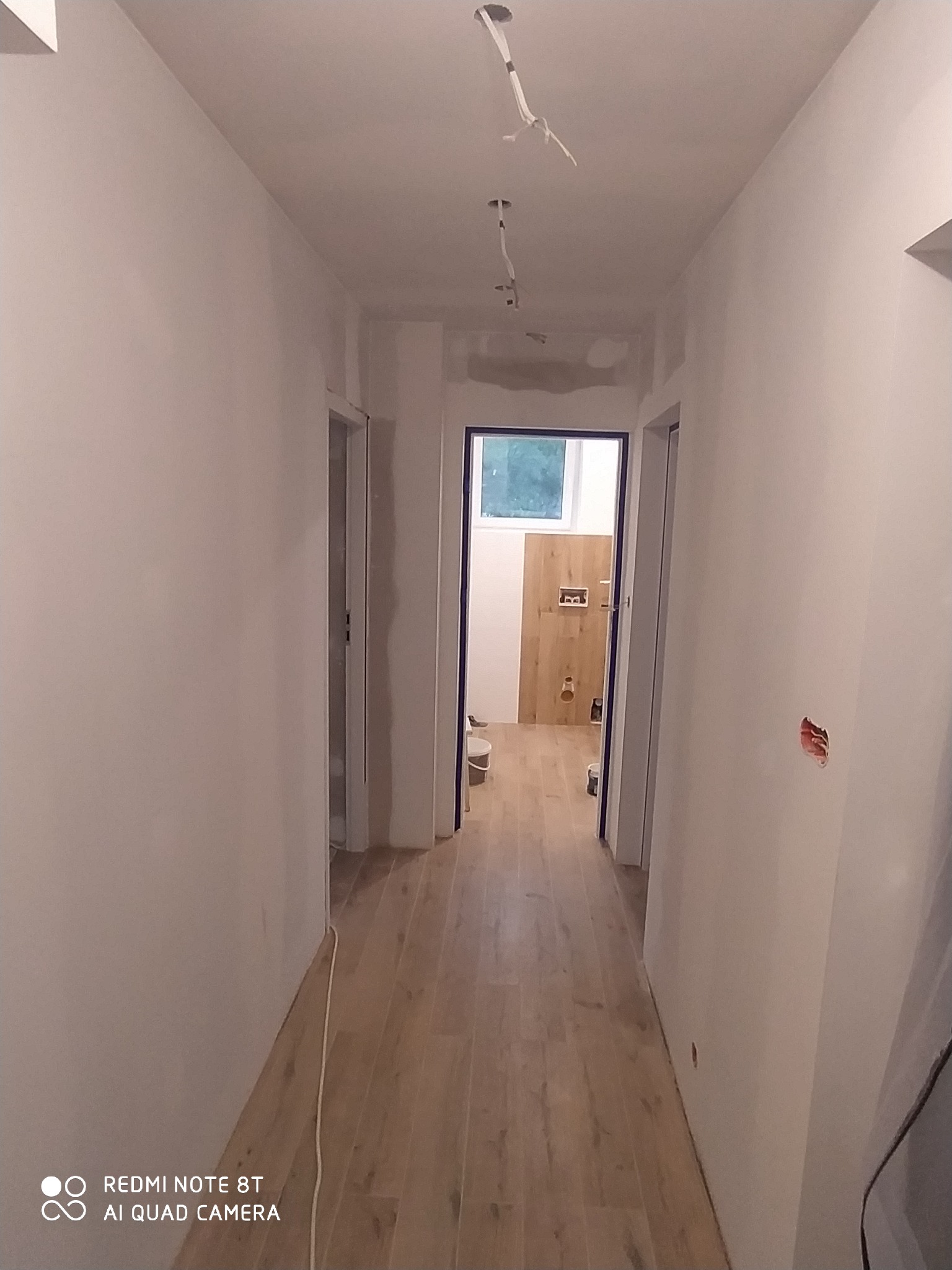 Korytarz w trakcie malowania na biało, z widoczną instalacją elektryczną na suficie i fragmentem wykończonej łazienki w tle.