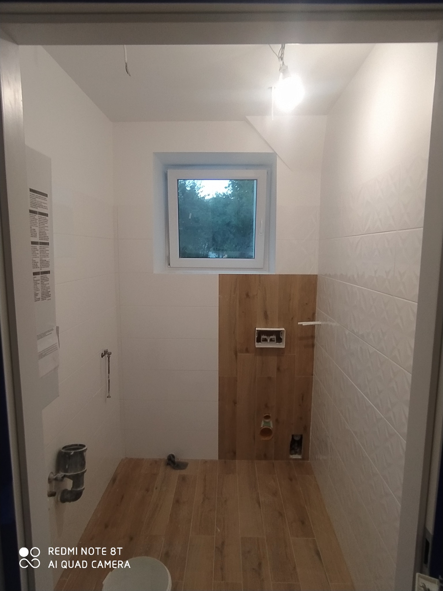 Wnętrze łazienki w trakcie remontu: białe płytki na ścianach, drewnopodobne panele na podłodze i częściowo na ścianie, widoczne instalacje wodno-kanalizacyjne, okno z widokiem na zieleń.