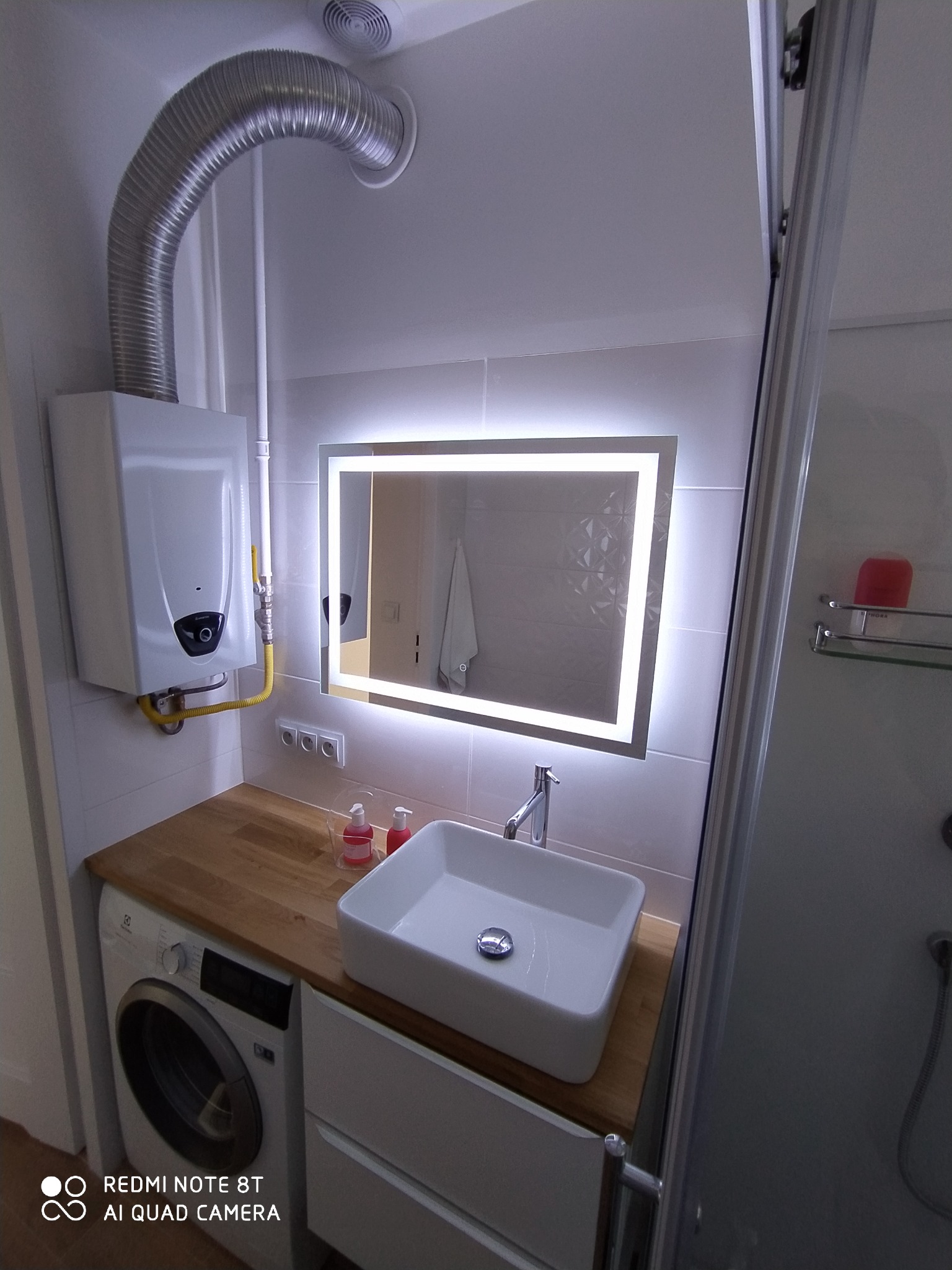 Nowoczesna łazienka z podświetlanym lustrem, białą umywalką nablatową, drewnianym blatem nad pralką i gazowym podgrzewaczem wody.