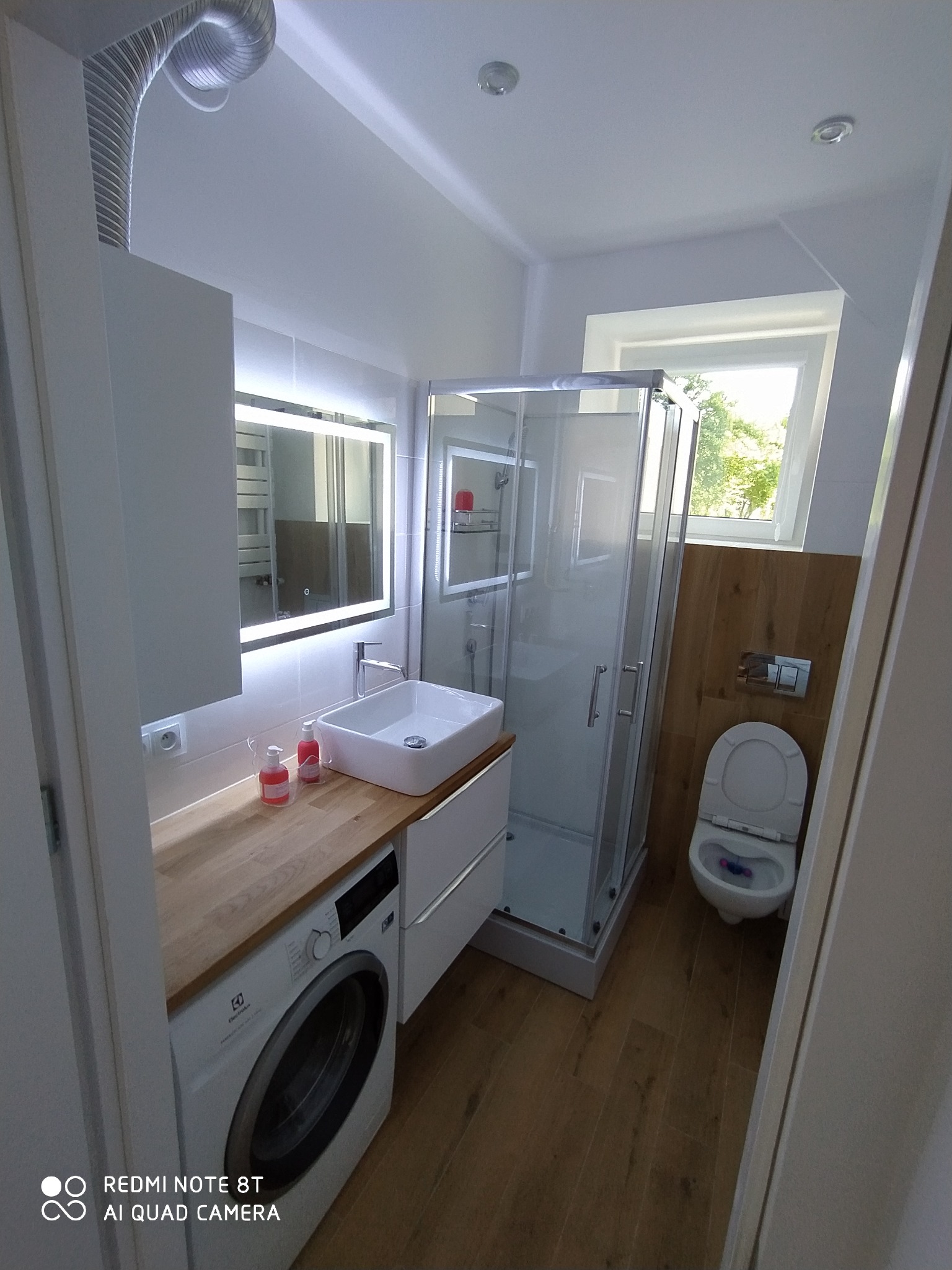 Nowoczesna łazienka z kabiną prysznicową, toaletą i umywalką nablatową na drewnianym blacie, pod którym znajduje się pralka; oświetlenie LED w lustrze.