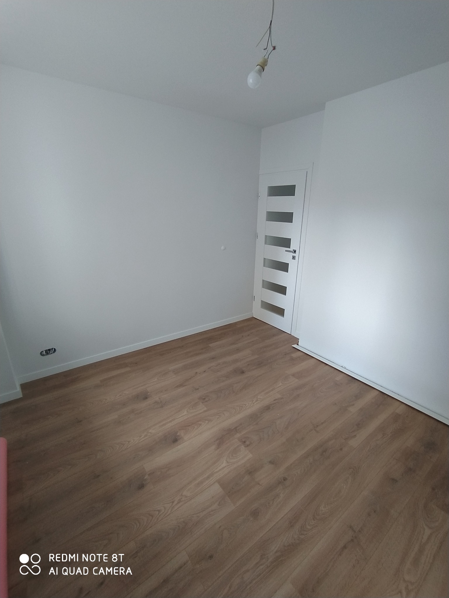 Pusty pokój z panelami podłogowymi w odcieniach brązu, świeżo pomalowane białe ściany i białe drzwi z przeszkleniami, surowa żarówka zwisająca z sufitu.
