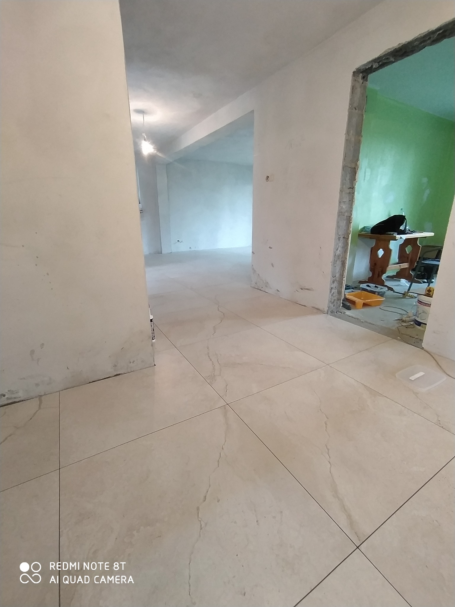 Beżowe, kwadratowe płytki podłogowe z marmurkowym wzorem po ułożeniu w trakcie remontu mieszkania, widoczne niedomalowane ściany i futryna drzwi, w tle pokój z pomalowaną na zielono ścianą.