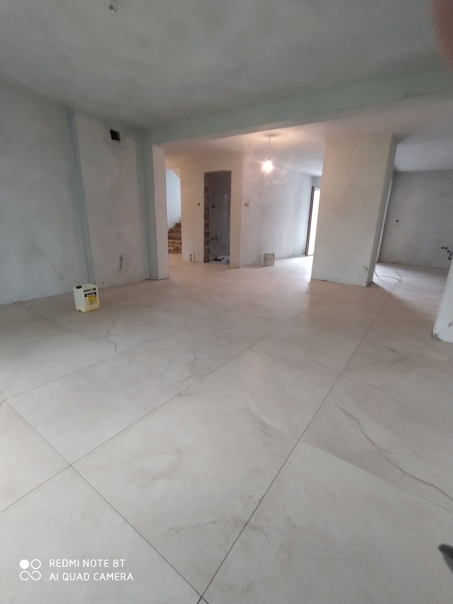 Wnętrze w trakcie prac wykończeniowych: jasne płytki podłogowe imitujące marmur, surowe ściany przygotowane do malowania, widoczna puszka z klejem do płytek w rogu pomieszczenia.