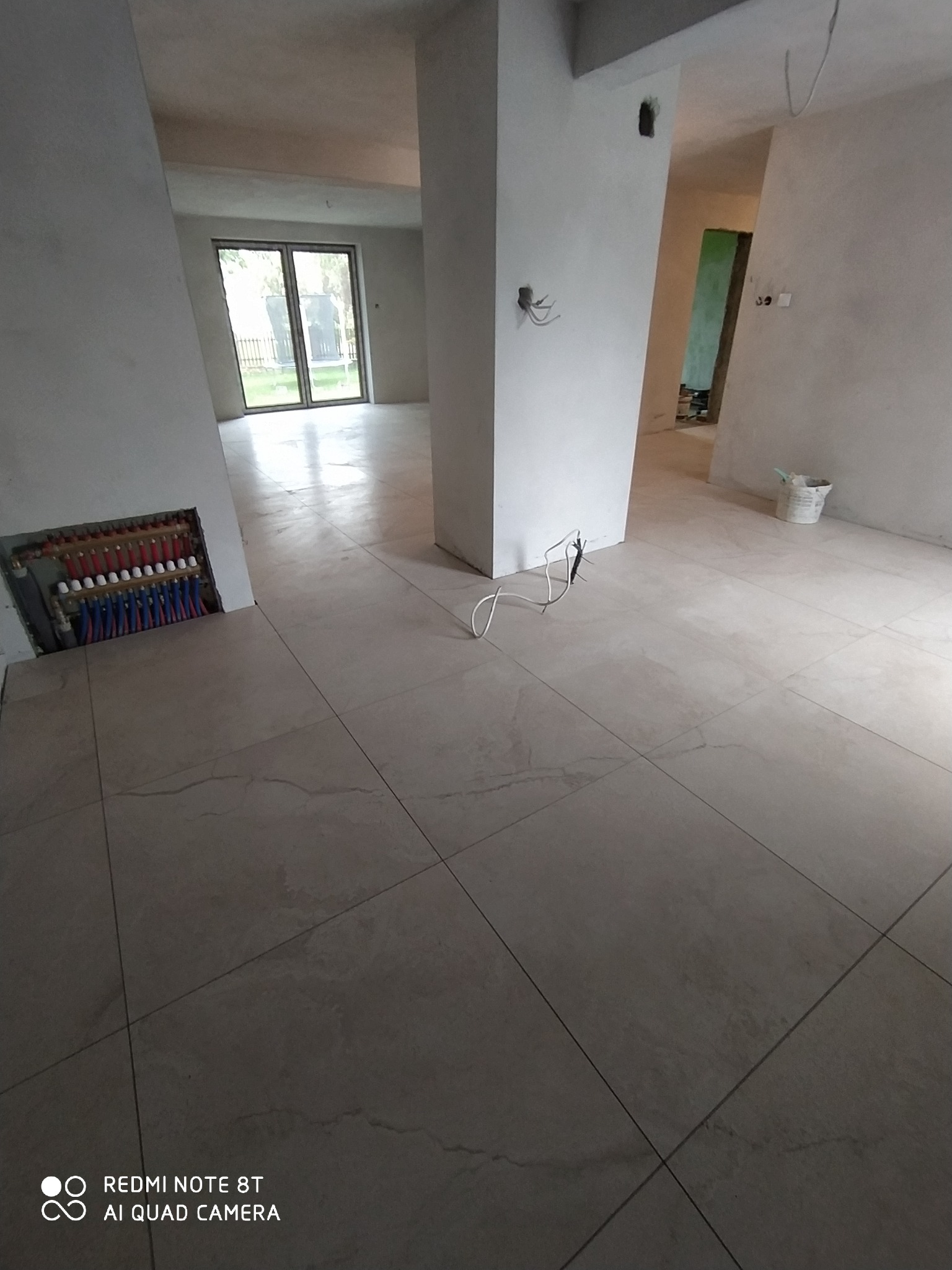 Wnętrze w trakcie remontu: Ułożone płytki podłogowe w odcieniach beżu z widocznym systemem ogrzewania podłogowego w ścianie, ściany przygotowane do malowania, widoczne przewody i puste puszki...