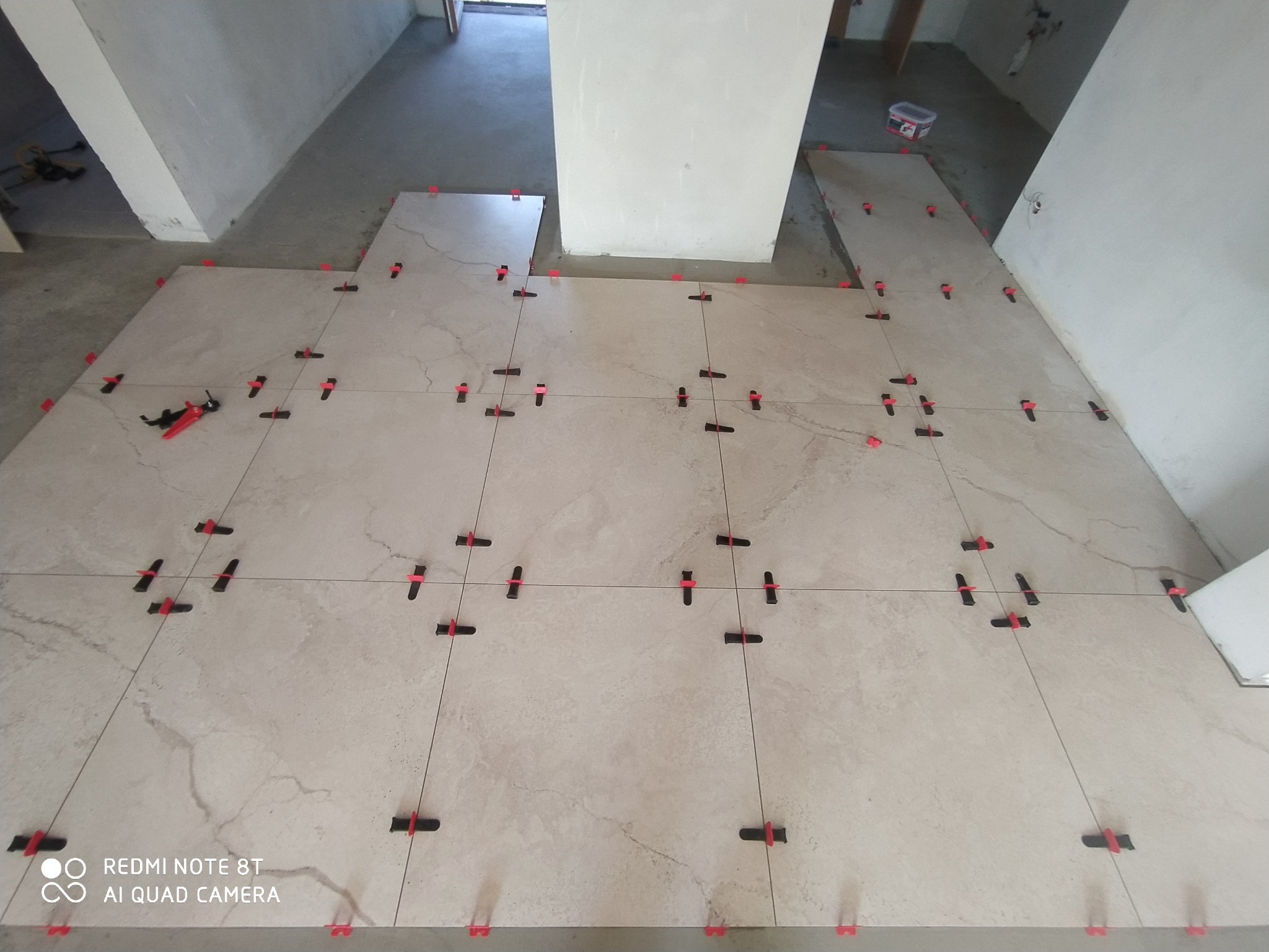 Jasne, beżowe płytki podłogowe w trakcie układania, z użyciem systemu poziomowania płytek (klipsy i kliny) oraz sznurka wyznaczającego linię, widoczne narzędzia glazurnicze w tle.