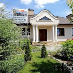 Jasny dom jednorodzinny z kolumnami przy wejściu, z brązowymi oknami i dachem, na tle zielonego ogrodu; na pierwszym planie rusztowanie z banerem reklamującym fotowoltaikę.