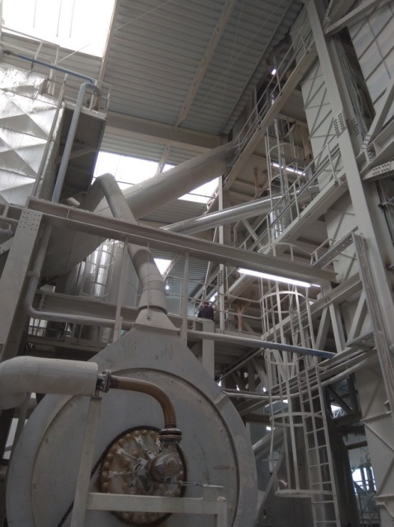 Wnętrze hali produkcyjnej z rozbudowaną instalacją przemysłową, widoczne rury, drabiny, zbiorniki i konstrukcja stalowa, zarys sylwetki pracownika na platformie.