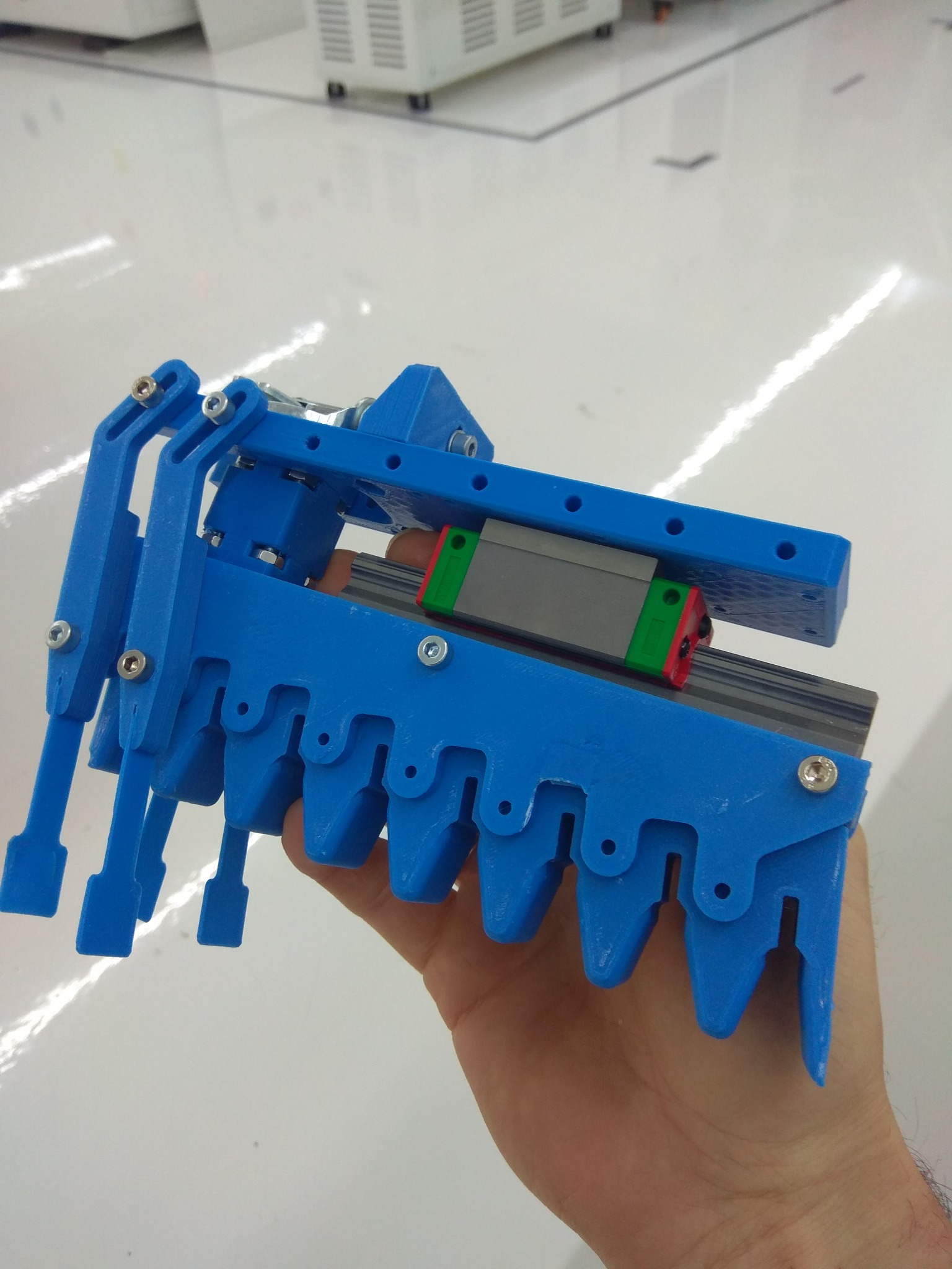 Niebieski, prototypowy element mechaniczny z tworzywa sztucznego, trzymany w dłoni, z widocznym prowadnikiem liniowym i elementami mocującymi.
