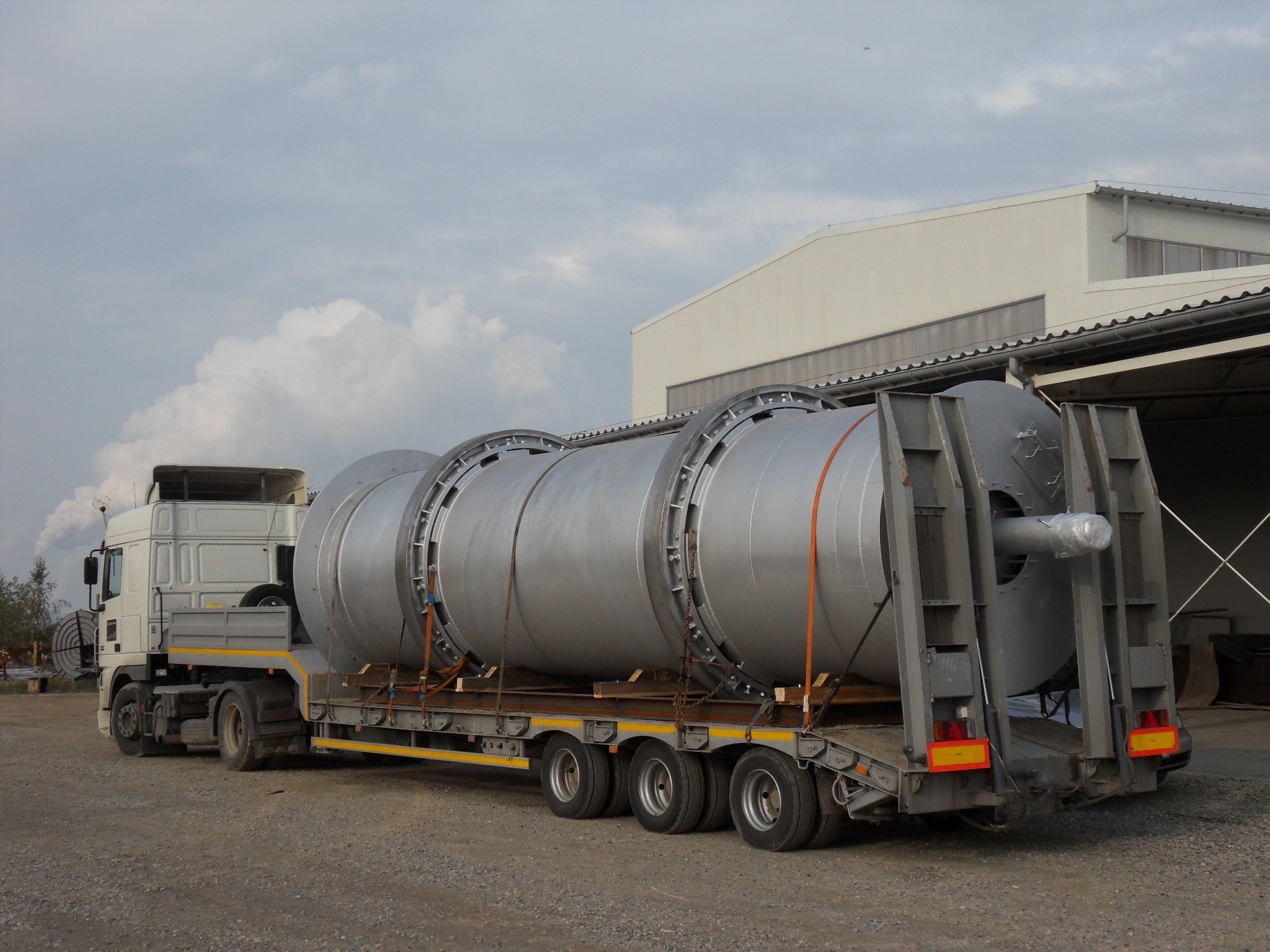 Ciężarówka transportuje duży, srebrny, cylindryczny zbiornik z elementami konstrukcyjnymi na naczepie, widoczny budynek przemysłowy w tle.