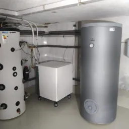 Instalacja kotłowni z dwoma zbiornikami buforowymi: biały z czarnymi otworami rewizyjnymi i szary, obok biały zbiornik wyrównawczy, rury izolowane na ścianie.