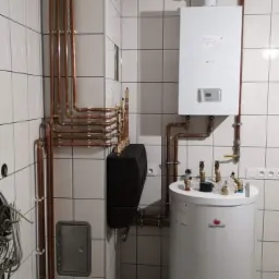 Kotłownia z piecem gazowym, zasobnikiem ciepłej wody i rozbudowaną instalacją miedzianą na tle białych płytek ceramicznych.