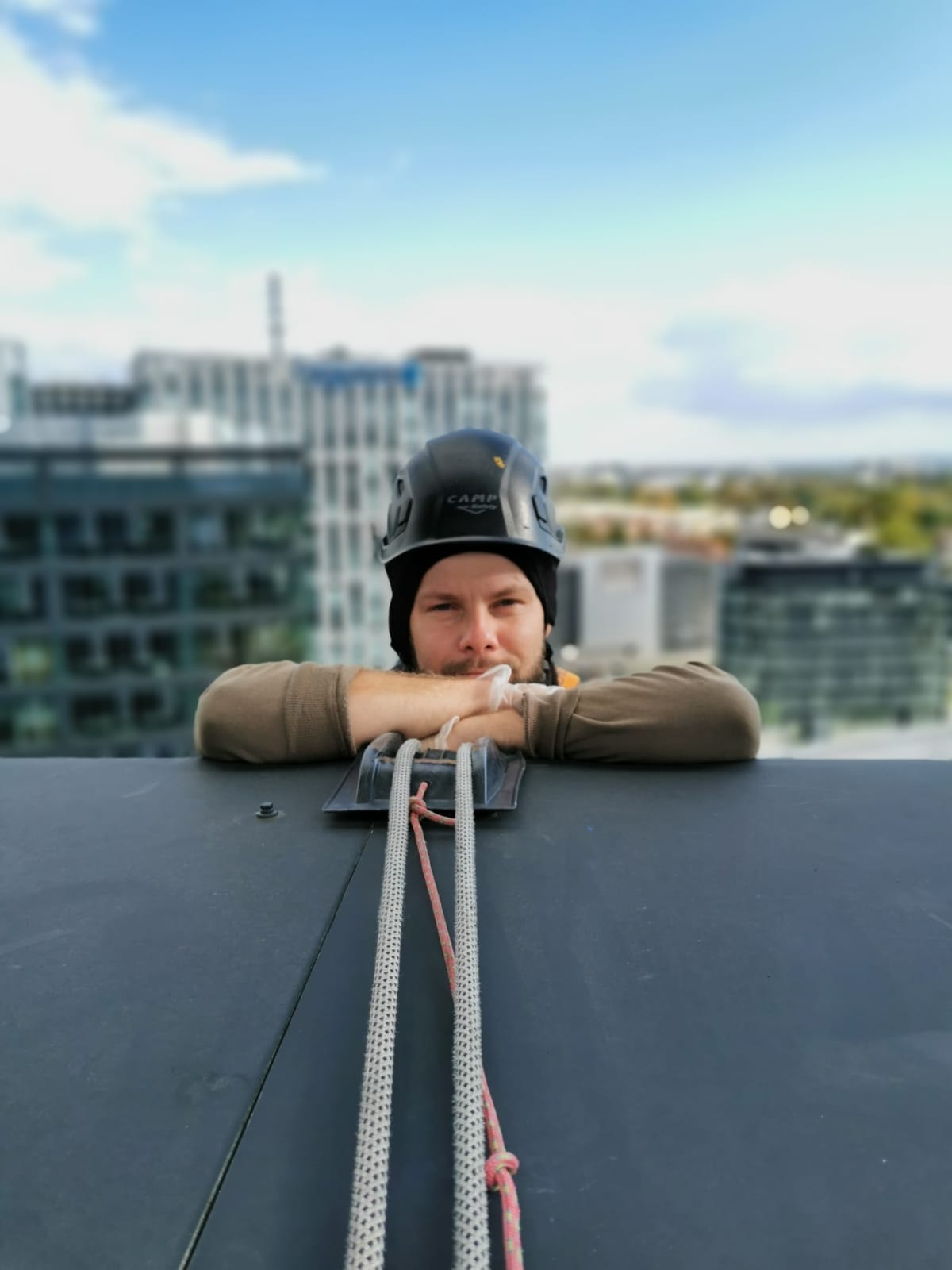 Pracownik w kasku CAMP opiera się o zabezpieczenie linowe na dachu wysokiego budynku, w tle widoczne inne budynki i niebieskie niebo.