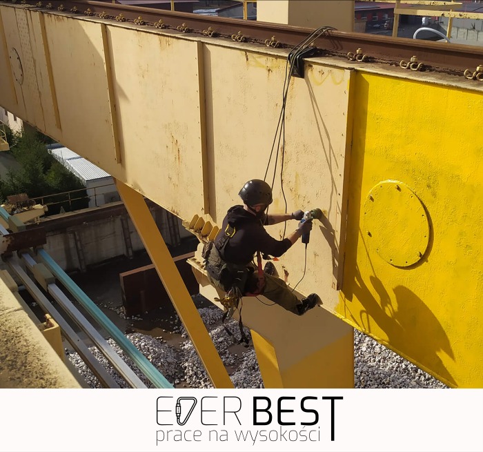 Pracownik w uprzęży, zawieszony na żółtej konstrukcji stalowej, wierci otwór w metalu, widoczne tory kolejowe na górze.