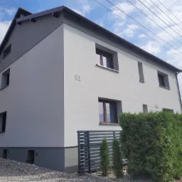 Dwukondygnacyjny dom jednorodzinny po remoncie elewacji, pomalowany na biało-szaro, z numerem domu '61' i brązowymi oknami, widok z zewnątrz na tle błękitnego nieba i zielonych tuj.