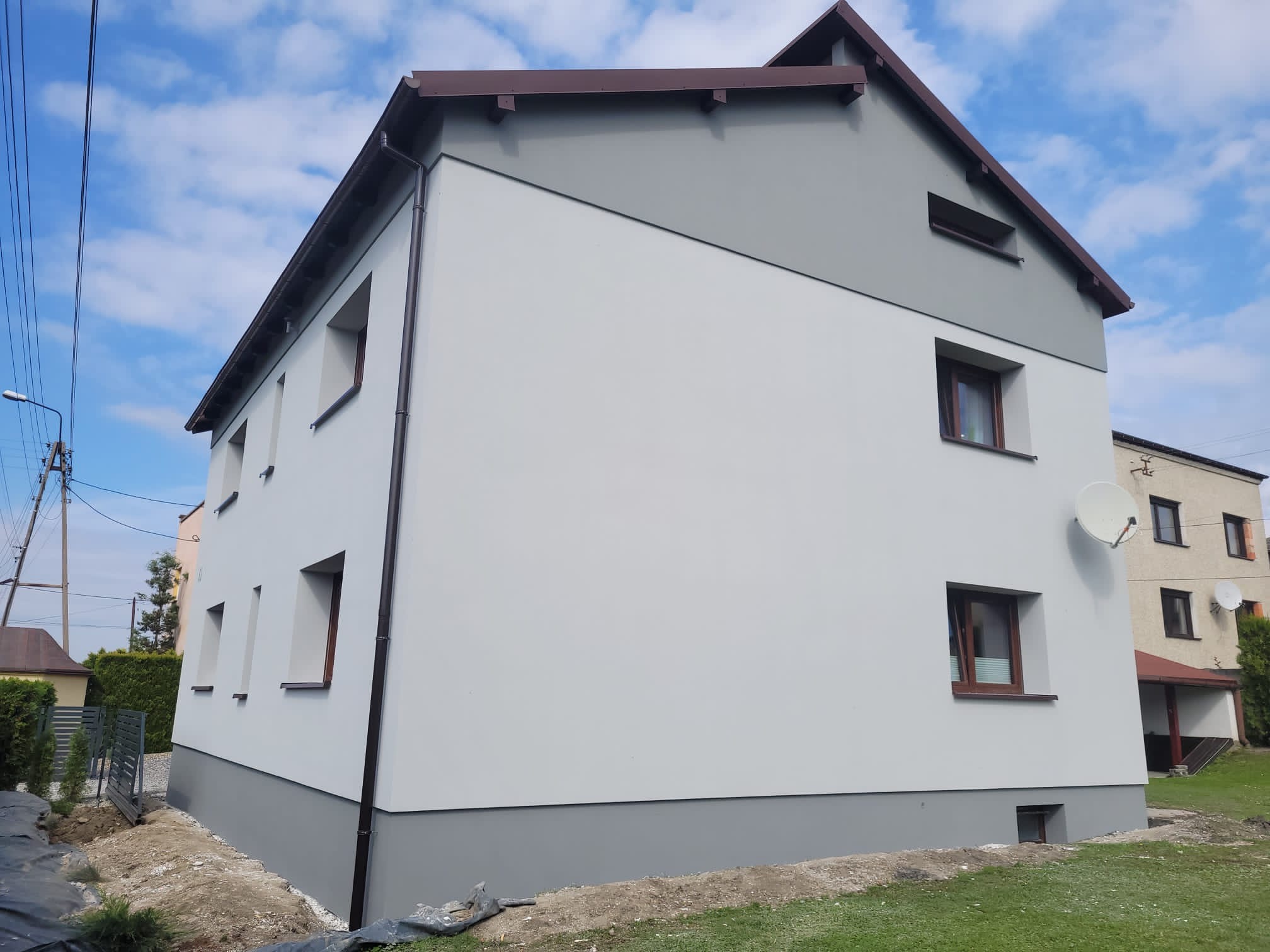 Dwukondygnacyjny dom po odnowieniu elewacji w odcieniach szarości i bieli, z brązowymi ramami okiennymi i dachem, na tle błękitnego nieba z chmurami.