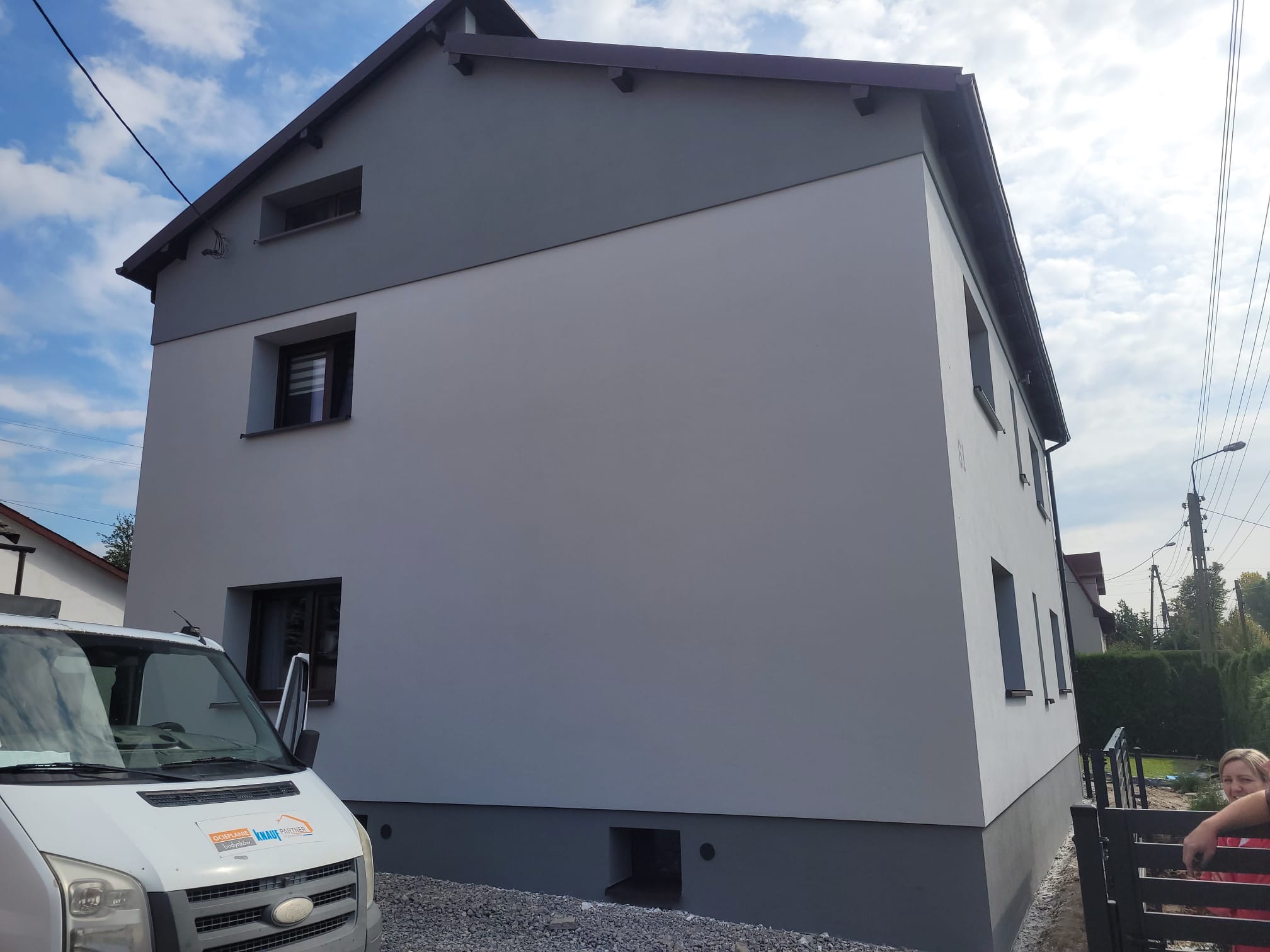 Dwukondygnacyjny dom z elewacją w odcieniach szarości, widoczny samochód dostawczy z logo firmy budowlanej i częściowo widoczna osoba przy ogrodzeniu.