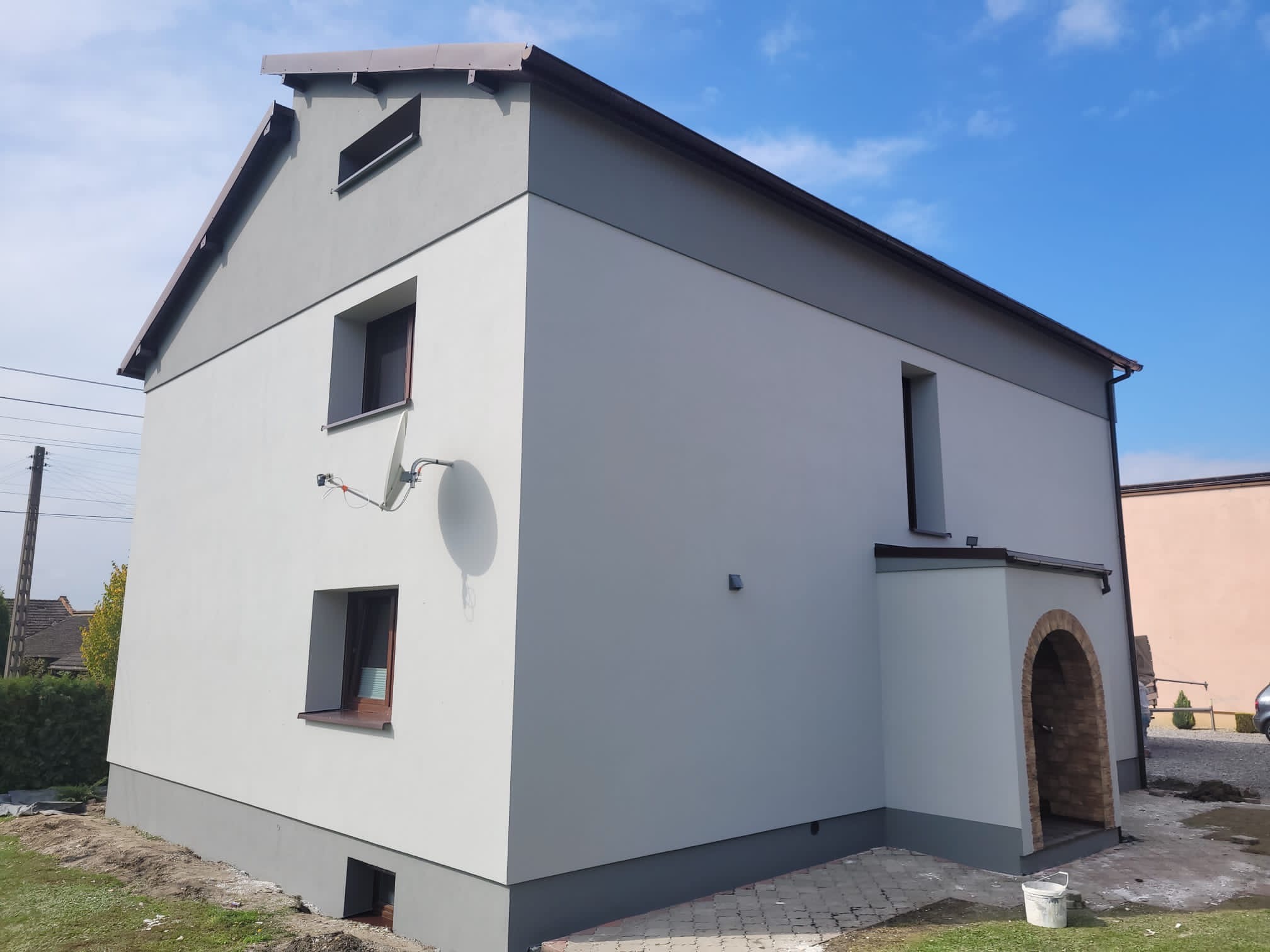 Dwukondygnacyjny dom z nową, dwukolorową elewacją w odcieniach szarości i bieli, z widocznym wejściem z ceglanym łukiem i anteną satelitarną na ścianie.