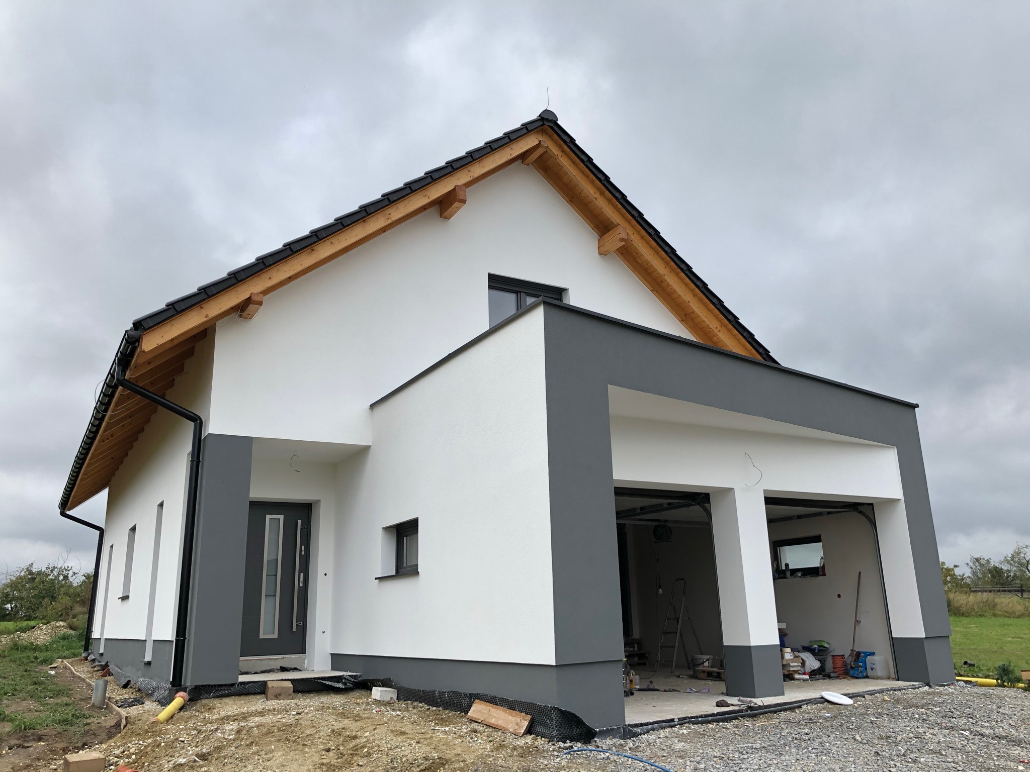 Nowoczesny dom jednorodzinny z garażem dwustanowiskowym, wykończony w bieli i szarości, widoczny na tle pochmurnego nieba. Elewacja świeżo pomalowana, wokół domu teren jeszcze niezagospodarowany.